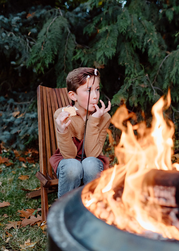 A boy who is enjoying leisure time outside with his family licks marshmallow off his fingers while eating a s'more he made with a marshmallow he roasted over a bonfire.