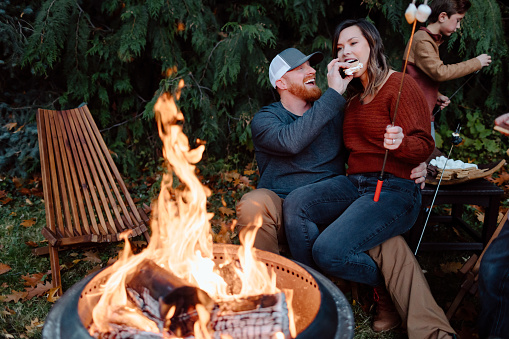 A woman sits on her husband's lap and takes a bite of a s'more while roasting marshmallows with their kids outside in the backyard.