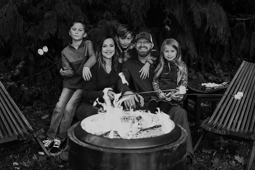 Black and white portrait of a husband and wife posing affectionately with their three elementary age children next to a fire pit while roasting marshmallows in the backyard of their home. All five individuals are smiling directly at the camera.