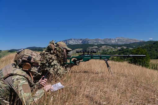 Sniper soldier assisted by an assistant to observe the area to be targeted with modern warfare tactical virtual reality goggles aerial drone military technology