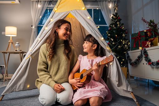 Caucasian mother playing with young daughter in decorated tent at home. Happy family, Beautiful mom teaching adorble kid girl using ukulele and singing a song in living room in house during Christmas.