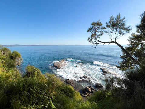 Horizontal high angle seascape photo of waves breaking on the rocky foreshore and grass and coastal scrub forest in Summer. Ulladulla, south coast NSW.