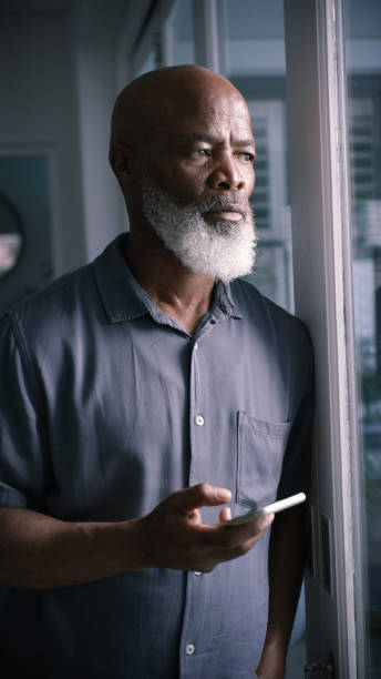 istock Serious, phone and communication with a senior black man thinking by a window in his home alone. Idea, focus and mad with a mature male standing by a glass door in his house during retirement 1451997301