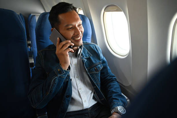 istock Smiling young hipster male passenger talking on mobile phone while sitting in airplane cabin near windows. 1452038771