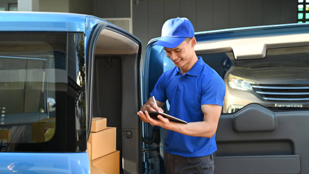 istock Delivery man in blue uniform using digital tablet next to open delivery van. Shipping and delivery service concept. 1452039571