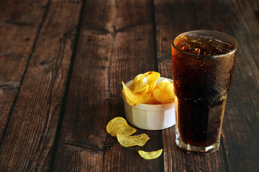 A glass of iced cola with potato chips on wooden table. Soft drinks and potato crisps.