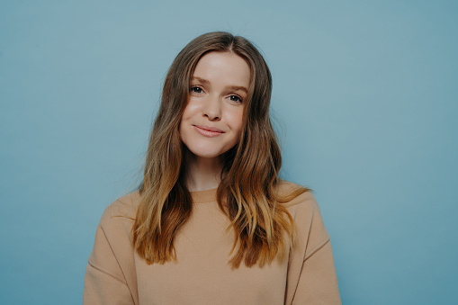 Photo of pretty smiling teenage girl without makeup looking at camera wearing comfortable light brown sweater posing isolated over blue blue studio background. Young people concept
