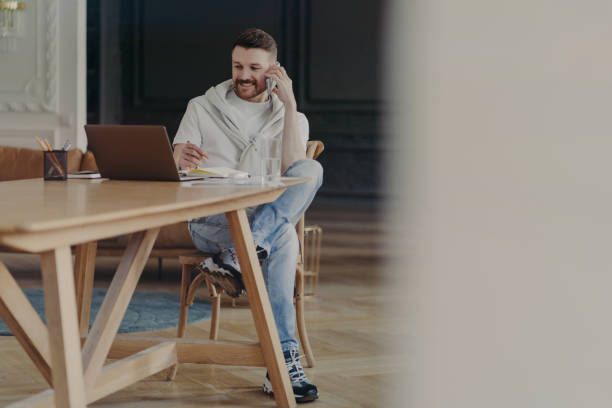 istock Young laughing man freelancer talking on mobile phone while sitting at his workplace at home, working remotely on laptop 1452098484