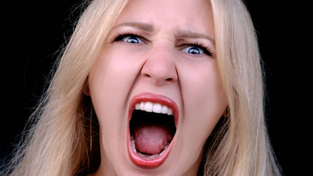 la donna arrabbiata urla di rabbia mentre guarda la telecamera su sfondo nero. headshot della ragazza che urla nella telecamera con la bocca aperta. risentimento, malcontento, odio. - mouth open women displeased anger foto e immagini stock