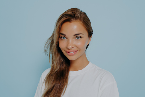 Close up portrait of smiling stylish young beautiful woman with long straight light brown hair in white t-shirt with natural light make up, isolated over blue studio background with copy space