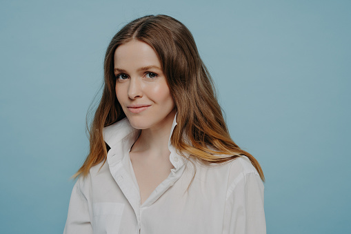 Confident beautiful teenage girl in white shirt with unbuttoned top being assured and looking at camera with slight smile while standing isolated on blue background, studio portrait