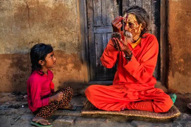 Little girl observing how Sadhu doing his makeup. In Hinduism, sadhu, or shadhu is a common term for a mystic, an ascetic, practitioner of yoga (yogi) and/or wandering monks. The sadhu is solely dedicated to achieving the fourth and final Hindu goal of life, moksha (liberation), through meditation and contemplation of Brahman. Sadhus often wear ochre-colored clothing, symbolizing renunciation.