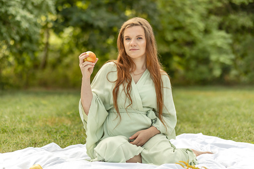 White pretty pregnant woman sits on blanket, holds,eats apple in park. Taking rest in meadow,relaxation. Healthy lifestyle. Baby delivery preparation,selfcare. Fresh air,trees on background.Horizontal
