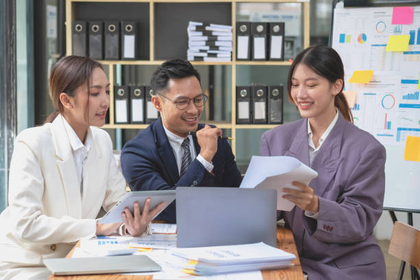 istock Team business three smiling and happy Asian men and women team using tablets and laptops brainstorm and plot statistics on a corporate chart board inside the company. Colleagues concept. 1452137532
