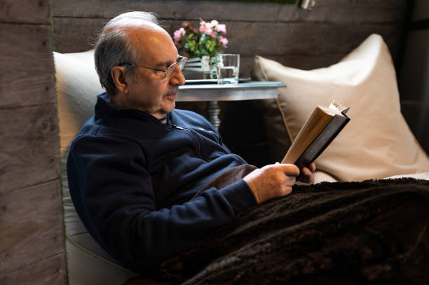 istock Elderly man with a mustache and reading glasses covered with a soft blanket sits on a bean bag and reads a book in cozy interior of wooden house 1452139679