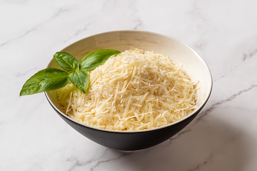 Grated parmesan in a bowl over marble background. Bowl of shredded grana padano cheese and green basil leaf prepared for cooking. Delicious italian hard cheese. Dairy product. Top view.