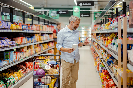 Mature male buyer shopping groceries in supermarket from shelf standing with shop cart