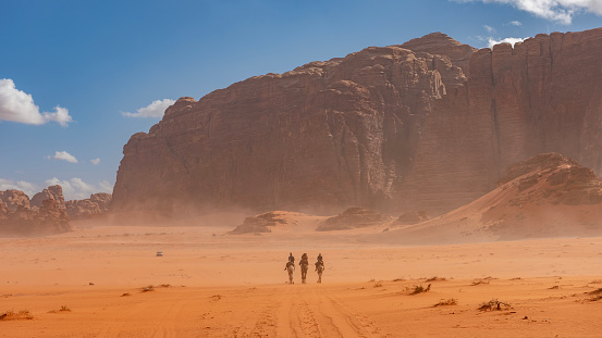 The Wadi Rum desert is one of the most picturesque and beautiful touristic spots of Jordan. In the picture three camel riders in the dust in front of a mountain ridge in the desert on a beautiful winter afternoon.