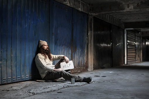 Old homeless man with gray beard wear sweater and blanket sitting food beggar because hungry and cold with holding metal glass seeking help from people walking pass on street. Help and hope concept.