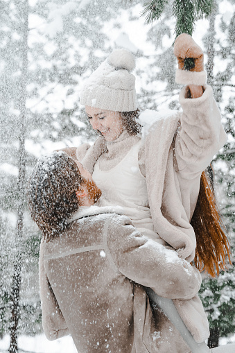Romantic snow love story.Young couple guy girl lying,playing in snowy winter forest with trees.Walking, having fun, laughing in stylish warm clothes, fur coat,woole jacket,shawl.Date,vacation weekend.