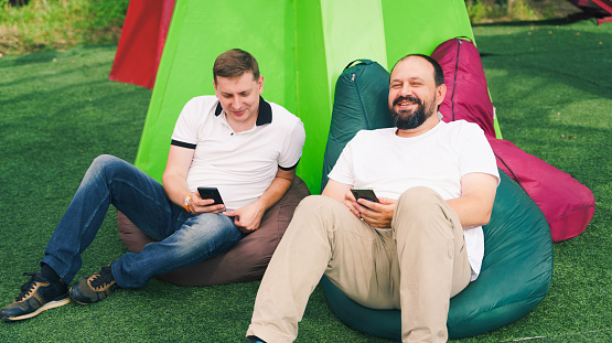 Two men,dads,friends sit on bean seat bags.Surfing in social networks in internet in mobile phone.Relax zone for parents on boring walks with children,kids.Playground in amusement park.Family weekend.