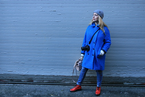 A Caucasian Polish woman in front of a blue wall. She is wearing a gray toque, blue coat, jeans, camera, a purse and red shoes.
