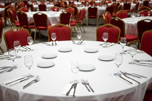 Tables ready for catering in the conference hall. White tablecloth, red chair, fork, spoon, knife, serving napkin, plate, glass.