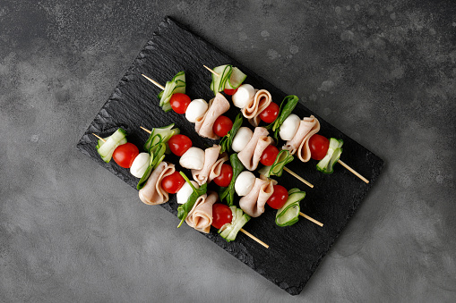 Mini appetizer on skewer Fresh Caprese Skewer. Antipasto, canape with meat, cucumber, Tomato and Mozzarella on dark slate board. Top view.