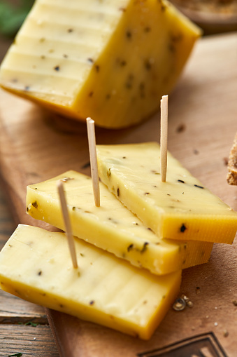 Cheese pieces on a wooden cutting board, on a kitchen table with a large piece of cheese
in the background, with bread crumbs, large copy space area