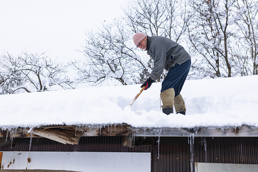 A senior man is on top of a farm house roof shoveling off snow.