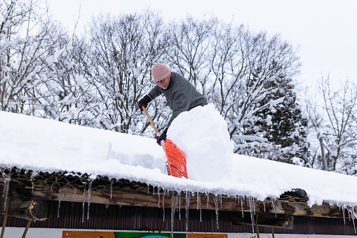 A senior man is on top of a farm house roof shoveling off snow.