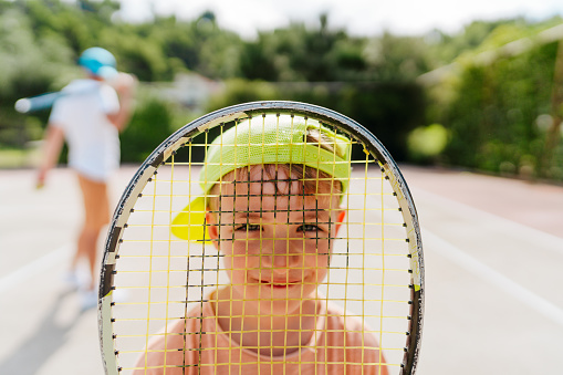 Photo of a little boy at the tennis court holding a tennis racket in front of his face