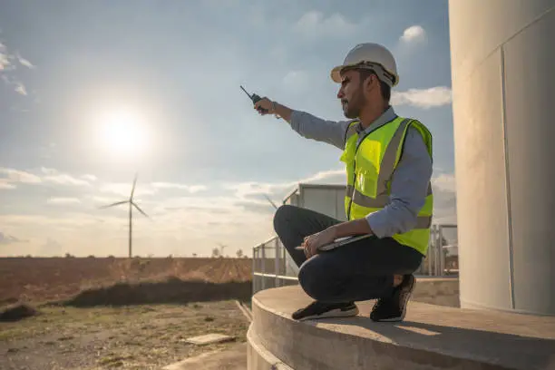 Asian engineer working in fieldwork outdoor. Worker sitting and inspect construction and machine around project site. Wind turbine electrical of clean resource energy  and environment sustainable.