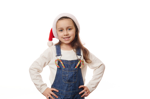 Beautiful Caucasian blue-eyed child, lovely little girl in Santa hat, smiling a cheerful toothy smile looking at camera, posing with Christmas lollipops over white background. Copy advertising space