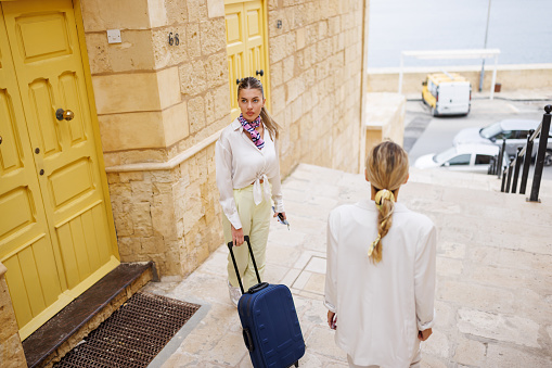 Young female friends with suitcase leaving their accommodation in old coastal town on sunny day