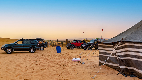 A group of cars parked by a tent in a bedouin camp in the Nafud desert of Saudi Arabia. In the background some camels.