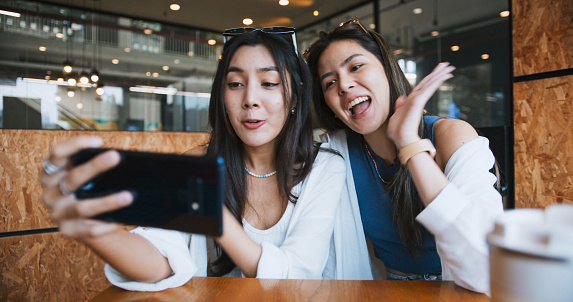 Young Asian female couple use mobile phone video call with friends, laugh and smile together at cafe. Smartphone internet communication information technology, remote meeting lifestyle concept