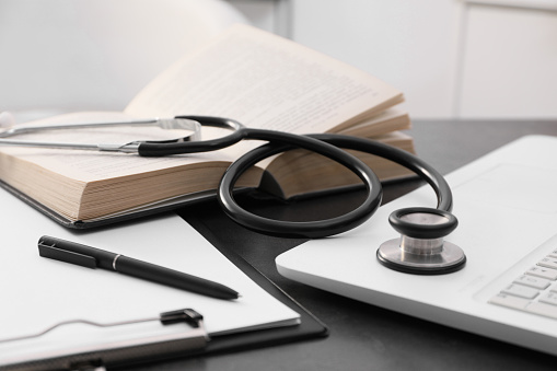Open student textbook, clipboard and stethoscope near laptop on grey table indoors, closeup. Medical education