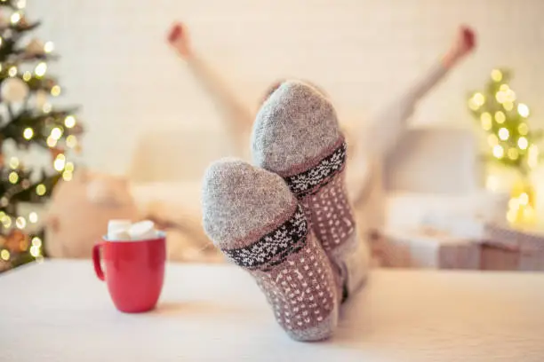 Photo of Woman sitting on couch near christmas tree. Female legs, feet in winter woolen socks putting on the table and relaxing after packing handmade  gift boxes. Presents for family.