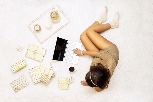 Christmas. Online shopping. Woman in sweater using laptop for making order, buying, lying on the white carpet among the many wrapped gifts in white gold wrapping paper. Top view. Concept