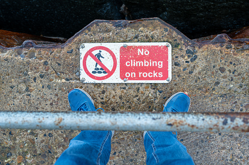 Shallow focus of a man standing at the edge of a ravine on a coastal path. A warning sign shows not to climb.