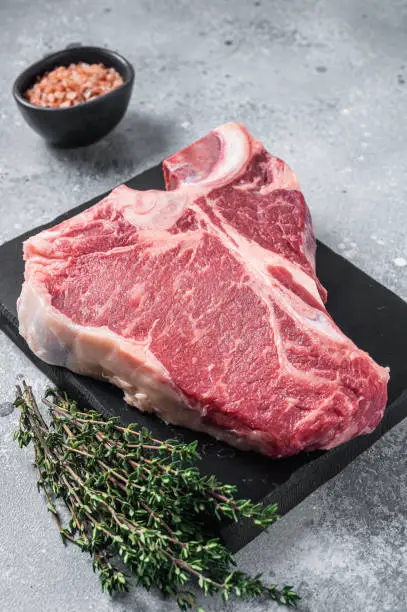 Raw dry aged wagyu porterhouse beef steak, uncooked T-bone on marble board with thyme. Gray background. Top view.