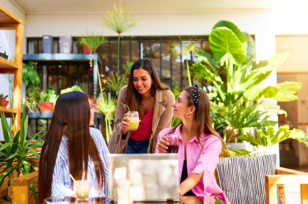 three young latin women sitting in front of a laptop talking smiling and drinking juice or coffee in a cafe stock photo