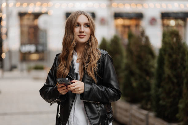 istock Beautiful smiling young blonde haired woman  in a black leather jacket using phone and making  video call on a city street background 1452339207