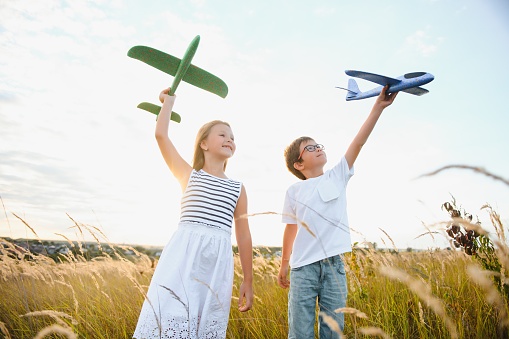 Running boy and girl holding two green and blue airplanes toy in the field during summer sunny day.
