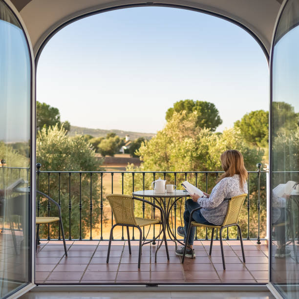 woman reading a book on the terrace of her room with large glass doors and views to the outside of a large garden. - resting place imagens e fotografias de stock