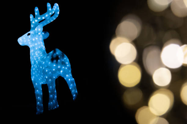 A close-up of a glowing candle inside a decorative lantern adorned with star patterns, creating a warm and magical Christmas ambiance. A blurred ceramic house in the background enhances the cozy holiday setting.