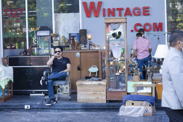 Market stall with  household objects at Vintage market stall opposite to Chatuchak Weekend market in Bangkok. Small vintage and second hand market outside of a vintage market shopping mall. A man wiuth sunglasses is sitting on  a chair. Other people are walking in scene