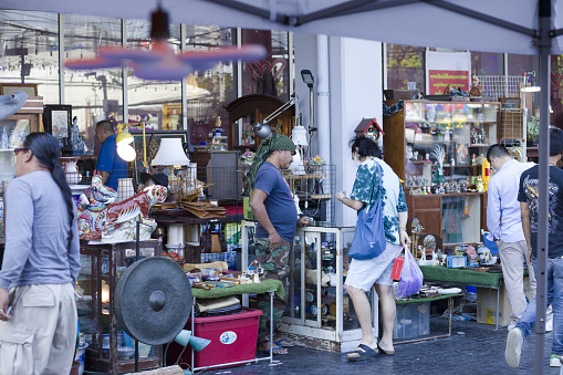 Tourist and thai vendor at  household objects Vintage market stall opposite to Chatuchak Weekend market in Bangkok. Small vintage and second hand market outside of a vintage market shopping mall. A man is is checking one item. Other people are around in scene