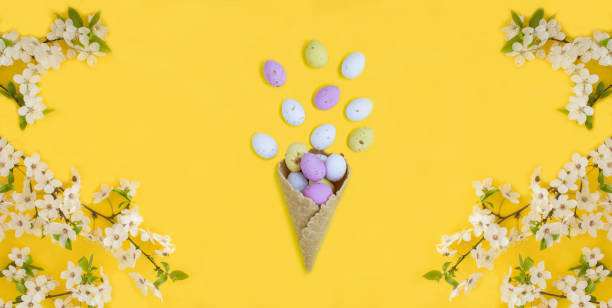 istock Ice cream cone with colored easter eggs and white flowering tree branches on the yellow background. Top view. 1452362223